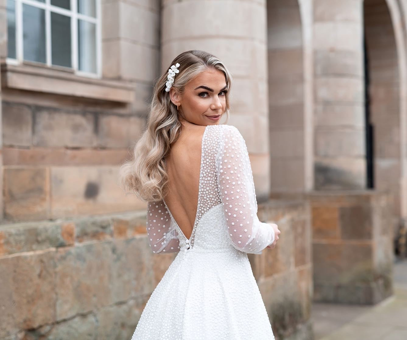 Blonde bridal model in front of historic building while looking over her shoulder while wearing Devoted bridal hair accessories positioned on right hand side of styled hair.