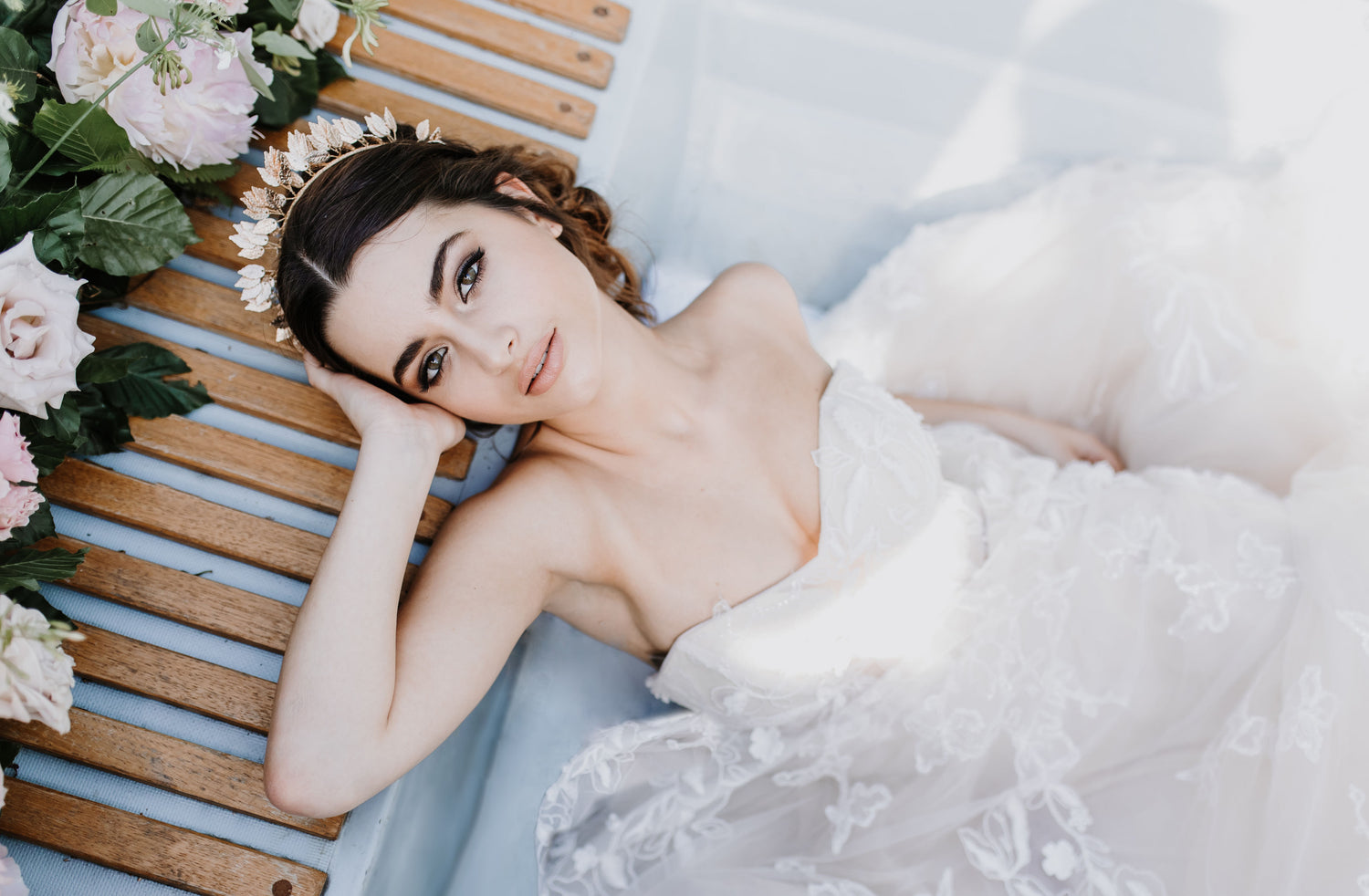 Dark haired model laying creatively with head positioned on slatted wooden base with flowers behind her head. All while wearing an ivory wedding dress and Devoted Gold Leaf Crown.